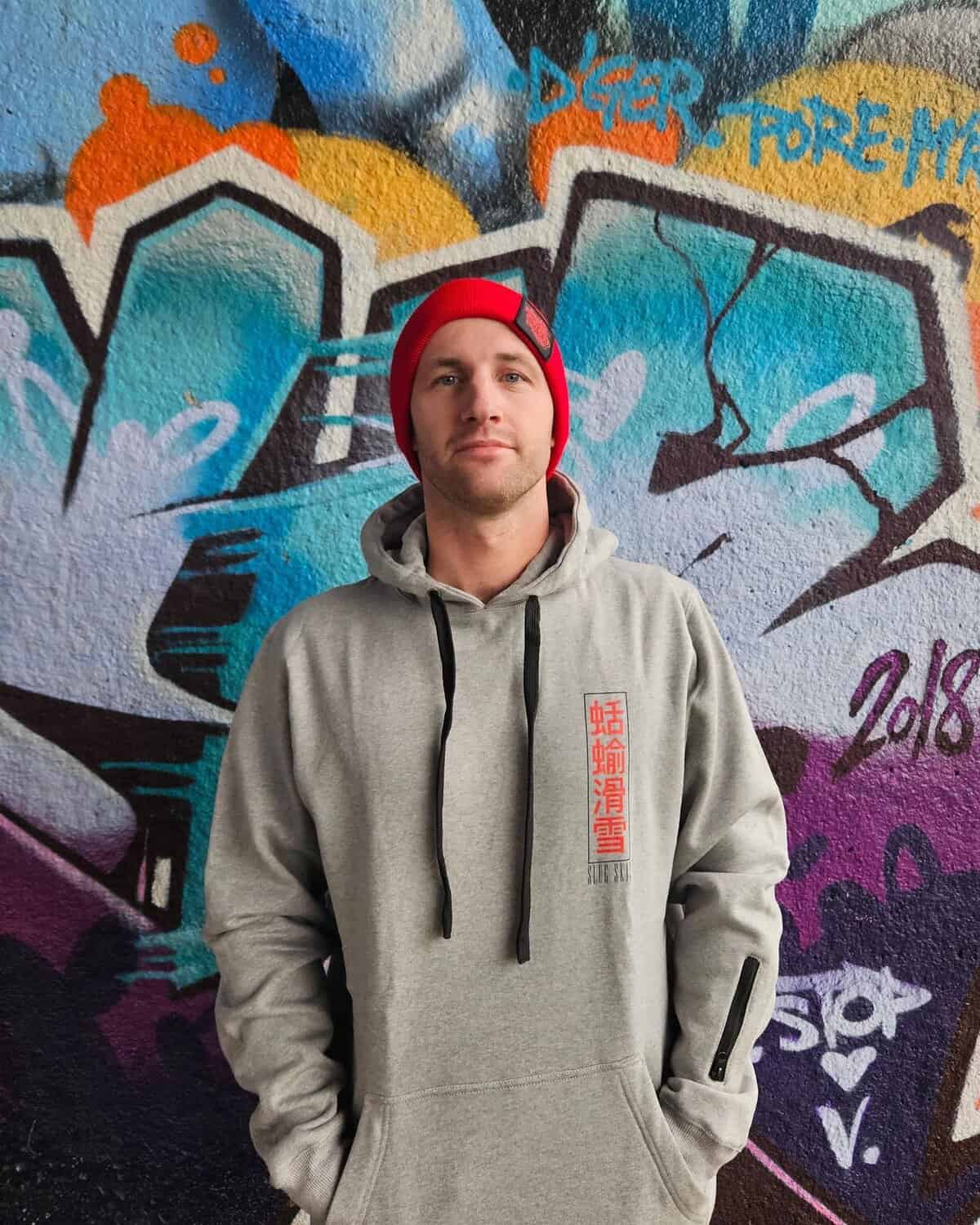 Alpine Hoodie Shinobu Grey Slug Skis Man Wearing Gear In Front Of Graffiti Wall